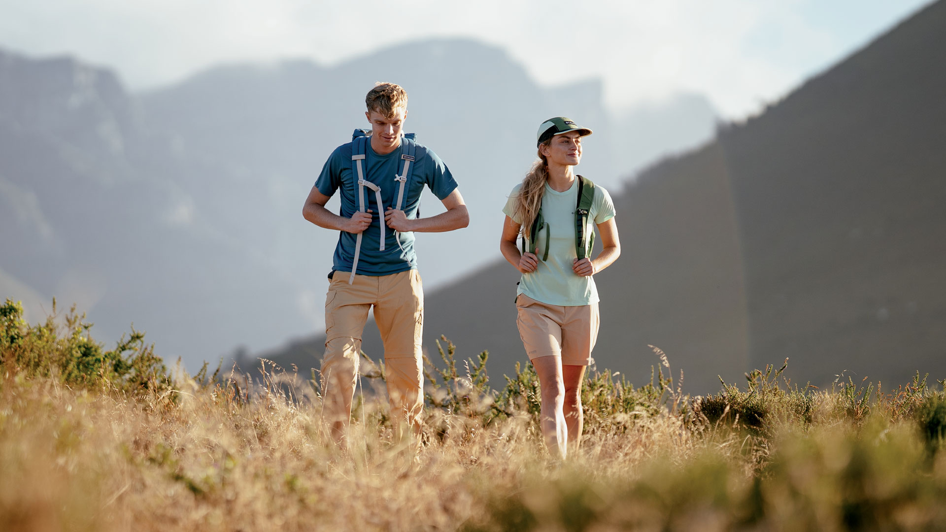 A man and woman in navy blue/black and purple/black respectively, hiking in brown boots, black backpacks on their back that have staps that go on their fronts, on the summit of a snow and rock capped mountain on a cloudy day. 