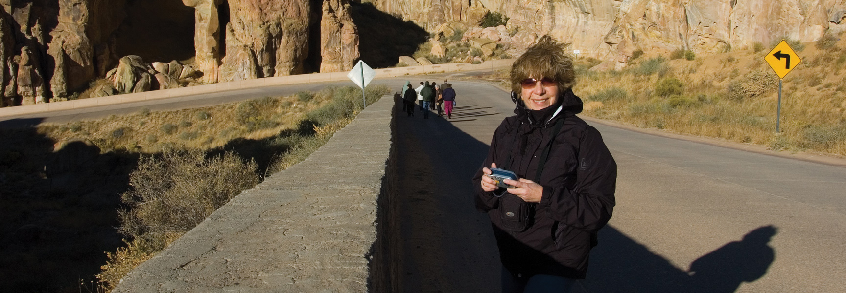 A woman smiles while holding a camera on a road in a rocky canyon near Albuquerque, with a tour group walking ahead.
