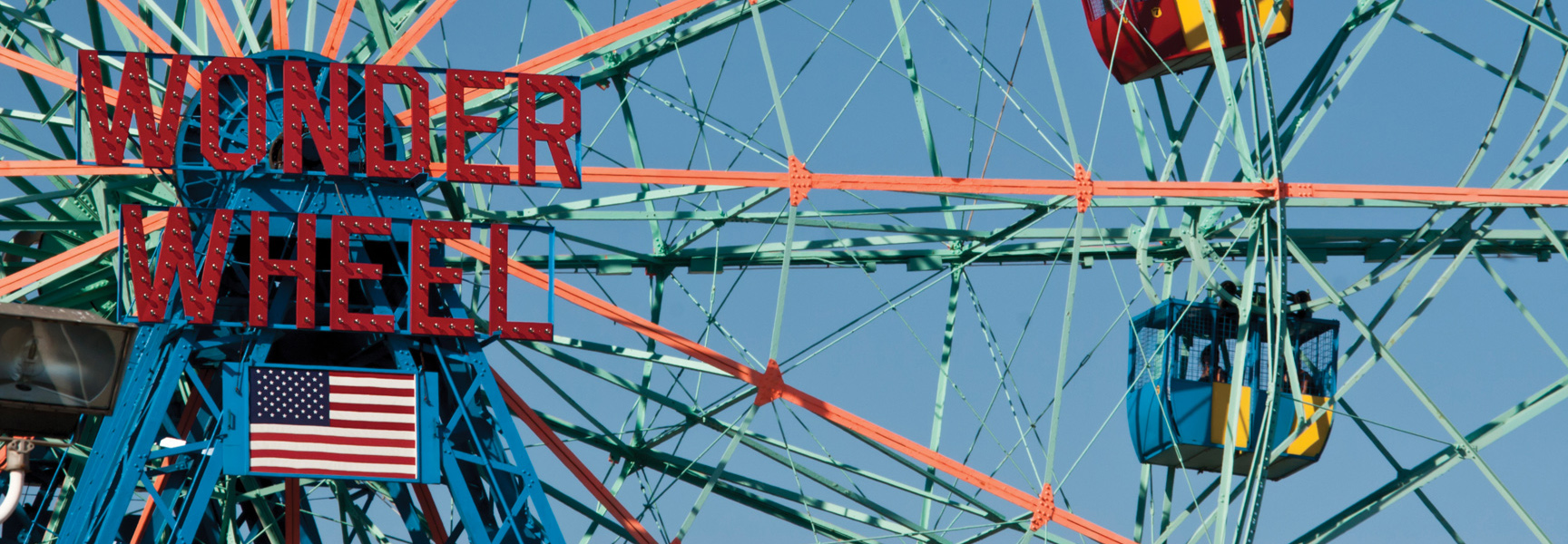 A close-up of the red Wonder Wheel sign and an American flag on the iconic ferris wheel in New York City against a blue sky.