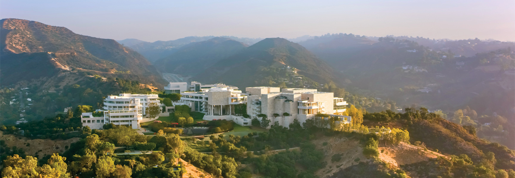 A sprawling modern museum with white stone architecture sits nestled in the hazy, rolling hills of Southern California at sunset.