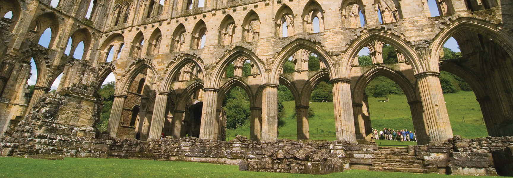 The sunlit ruins of a grand stone abbey in Britain, with rows of arches revealing a lush green lawn and rolling hills.