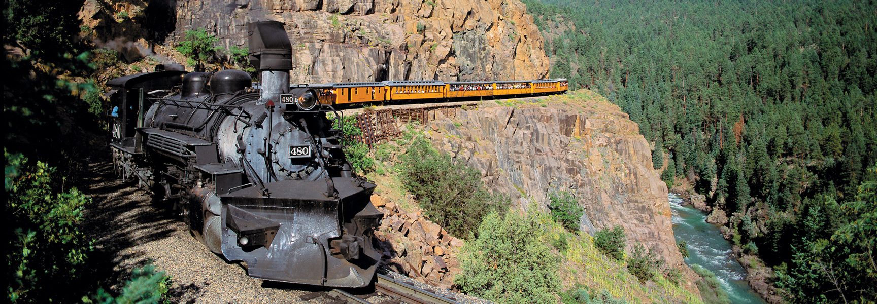A historic black steam engine pulls yellow passenger cars along a steep, rocky cliffside above a river in the mountains of Colorado.