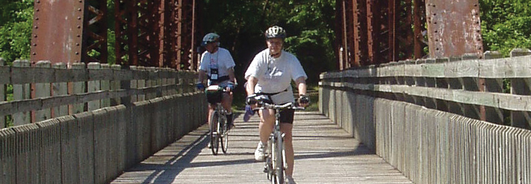 Two cyclists ride their bikes across a wooden bridge on the Katy Trail in Missouri, with a rusty metal trestle and green trees behind them.