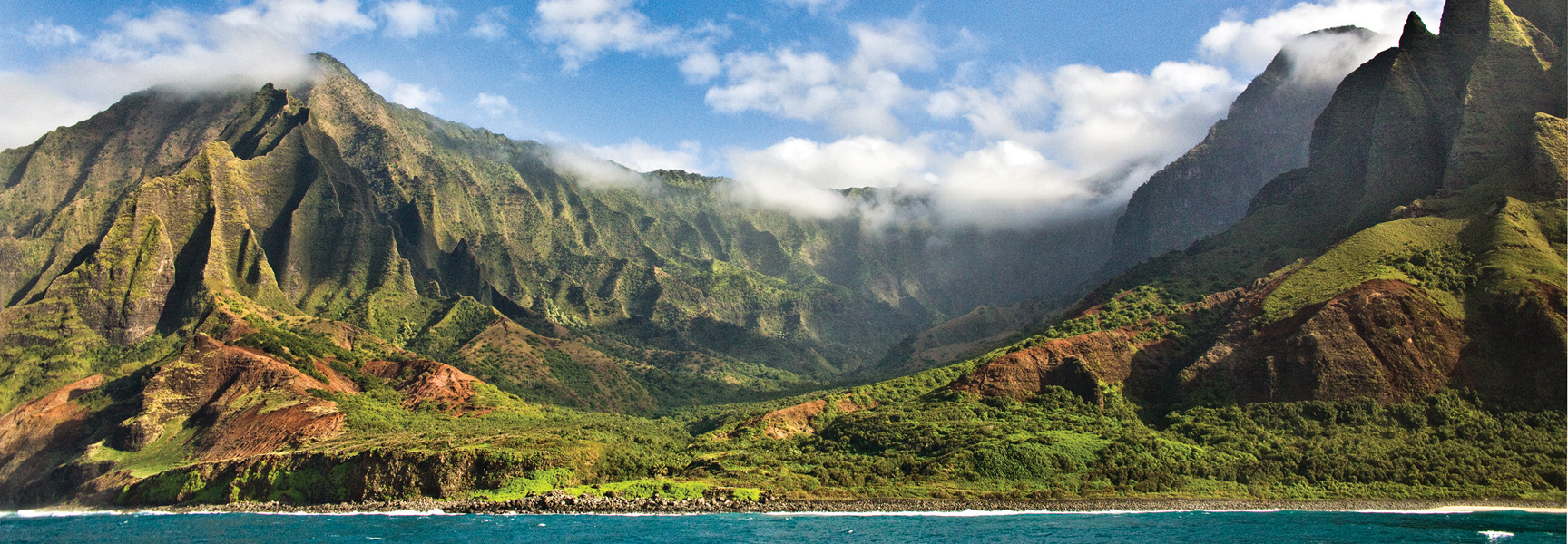 Lush green mountains with dramatic ridges rise above the blue ocean on the coast of Hawaii.