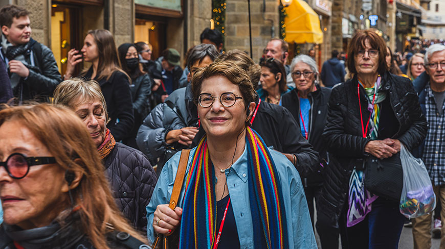 A woman with glasses and a colorful scarf smiles while walking with a tour group through a crowded street in Italy.