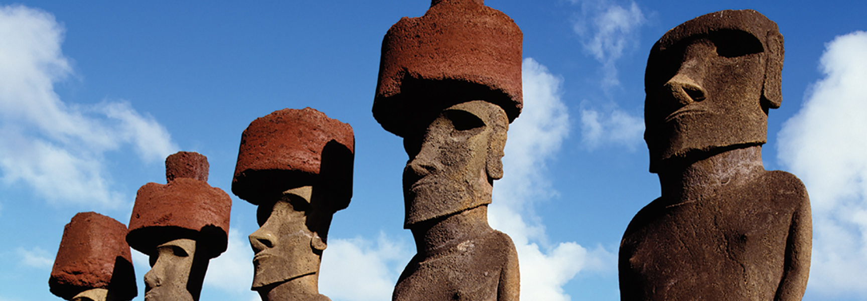 A row of ancient Moai statues with red topknots stand against a blue sky on Easter Island (Rapa Nui).