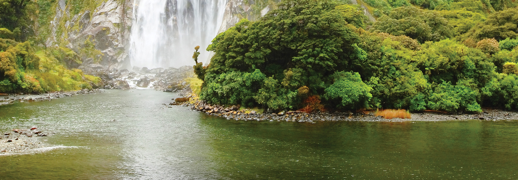 A wide waterfall cascades into a river surrounded by a lush, rocky, tree-covered landscape in New Zealand.
