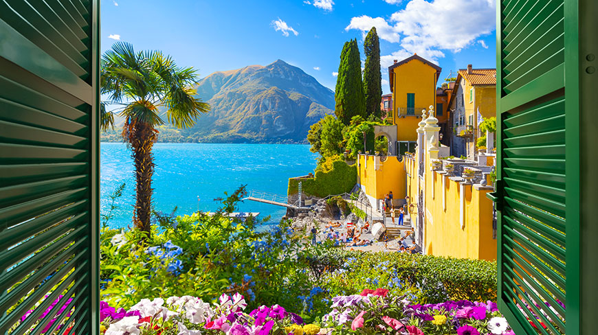 A view through green shutters of colorful flowers, yellow villas, and a bright blue Lake Como with mountains in Italy.