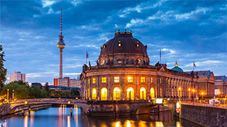 The illuminated Bode Museum at dusk in Berlin, Germany, with the Fernsehturm television tower rising in the background against a dramatic sky.
