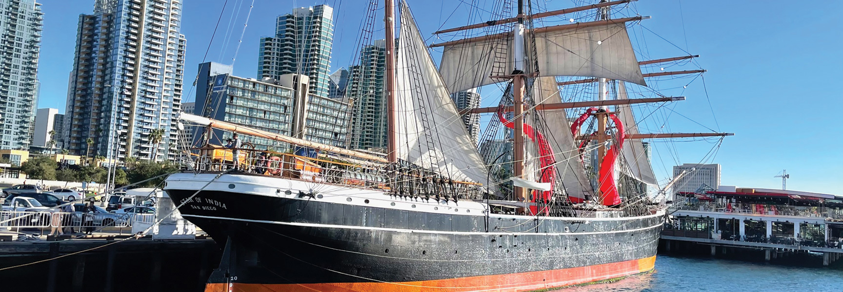 A tall ship named the Star of India is docked in the San Diego, California harbor with city skyscrapers in the background.