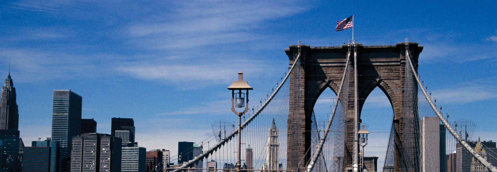 An American flag flies atop a tower of the Brooklyn Bridge, with the New York City skyline in the background.