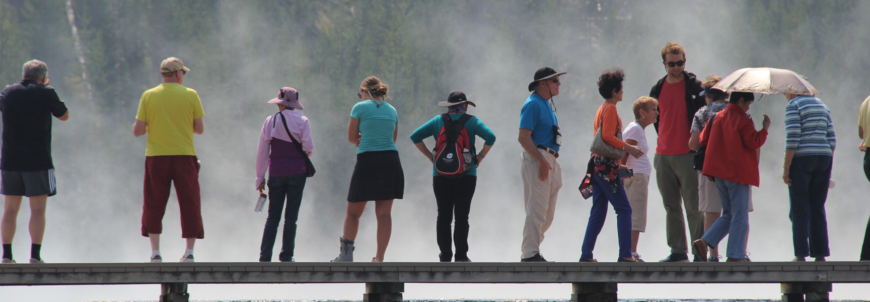 A group of visitors stand on a wooden boardwalk in Montana, watching steam rise from a geyser at Yellowstone National Park.