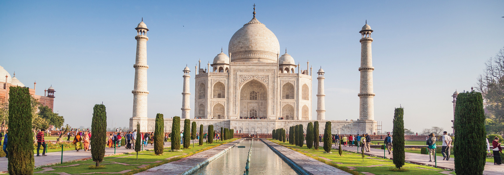 The white marble Taj Mahal in India, with its reflecting pool and gardens, is busy with tourists on a bright, sunny day.
