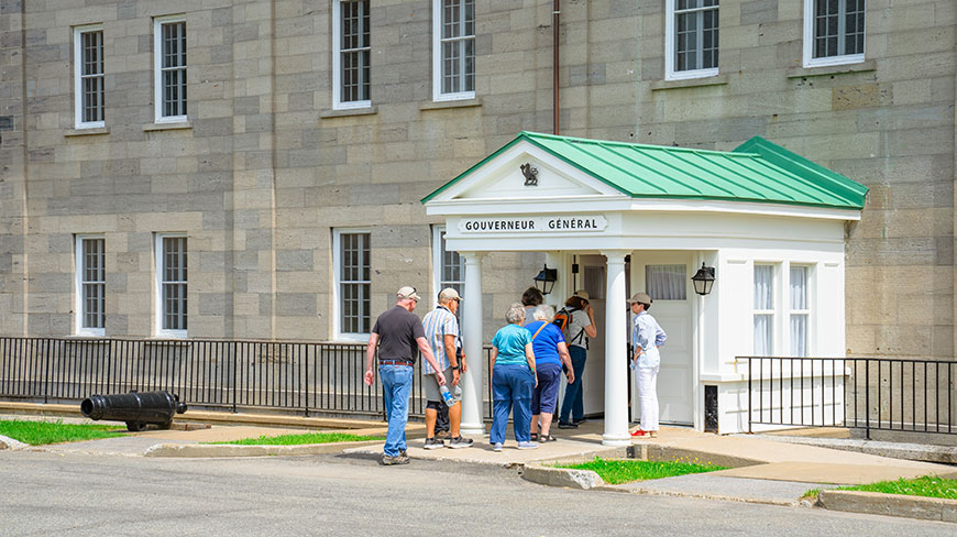 A group enters the stone La Citadelle Residence in Québec City through a white portico labeled Gouverneur Général next to a small cannon.