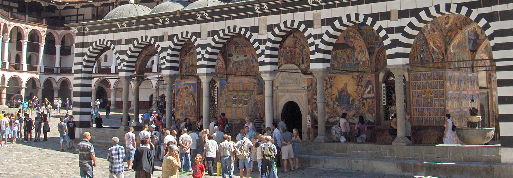 Tourists gather in the courtyard of the Rila Monastery in Bulgaria, admiring its striped arches and colorful frescoes on a sunny day.