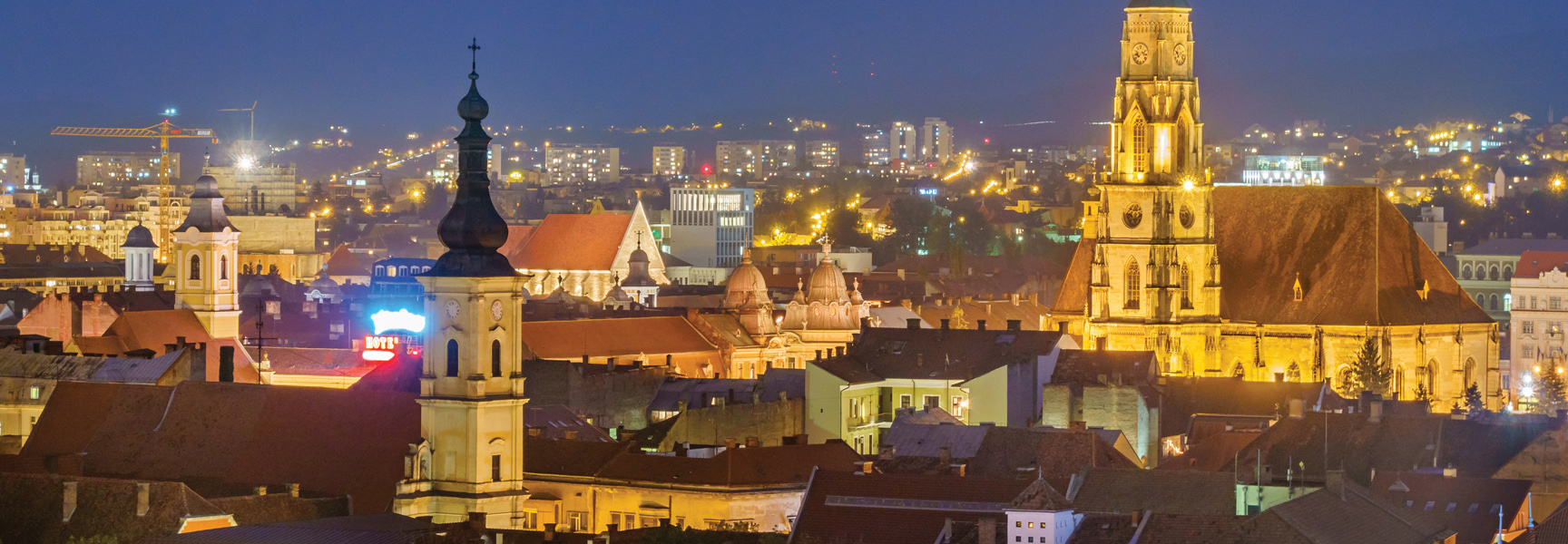 The illuminated historic steeples and city buildings of Cluj-Napoca, Romania are lit up against the twilight sky.