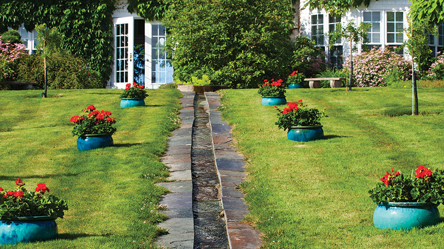 A stone rill runs through a green lawn dotted with bright blue pots of red flowers at Kingsbrae Garden in New Brunswick.