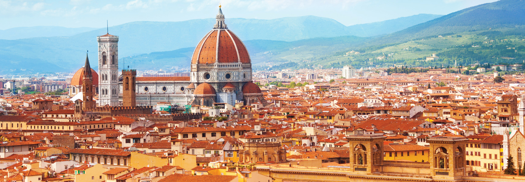 A scenic view of Florence, Italy, highlights the iconic Duomo and surrounding red-roofed buildings with mountains in the background.