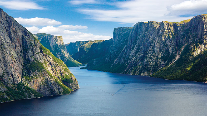 A boat sails through the deep blue waters of a majestic fjord, flanked by steep, green cliffs in Gros Morne, Newfoundland and Labrador.