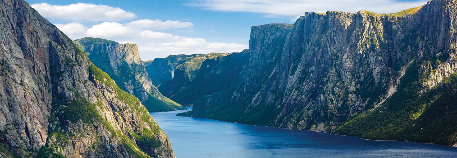Steep, rocky cliffs covered in green vegetation rise from a deep blue fjord in Gros Morne National Park, Newfoundland and Labrador.