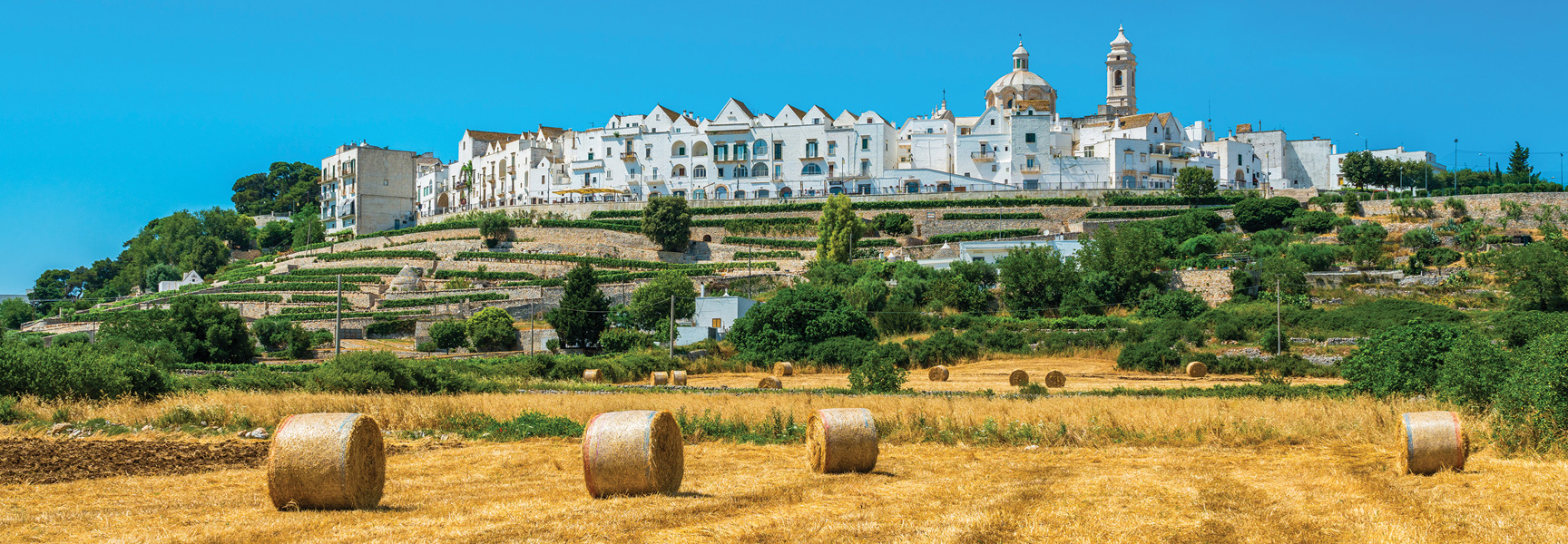A white-washed hilltop town in Southern Italy overlooks terraced vineyards and a golden field with large, round hay bales under a clear blue sky.