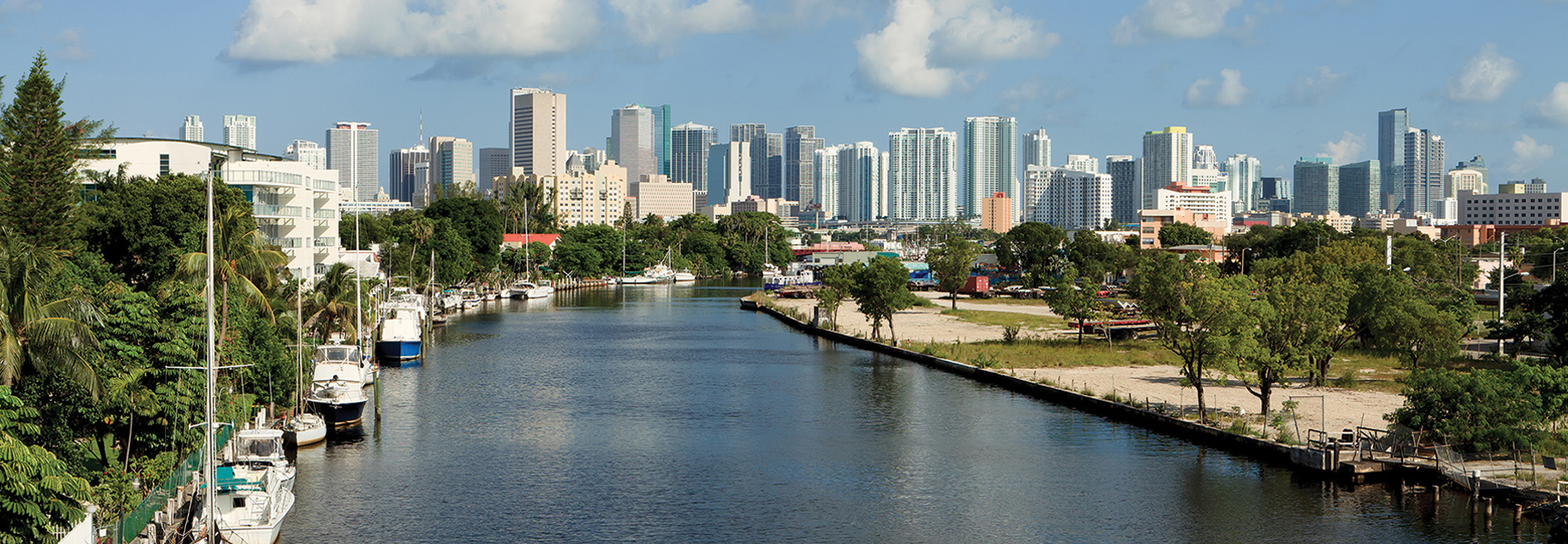 Boats are docked along the Miami River with the downtown Miami, Florida skyline visible in the background under a partly cloudy sky.
