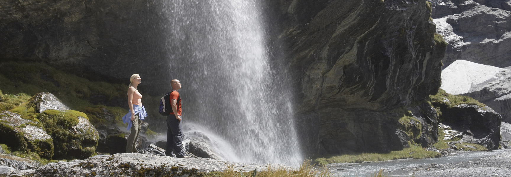 Two hikers stand on a rock and admire a large waterfall in New Zealand.