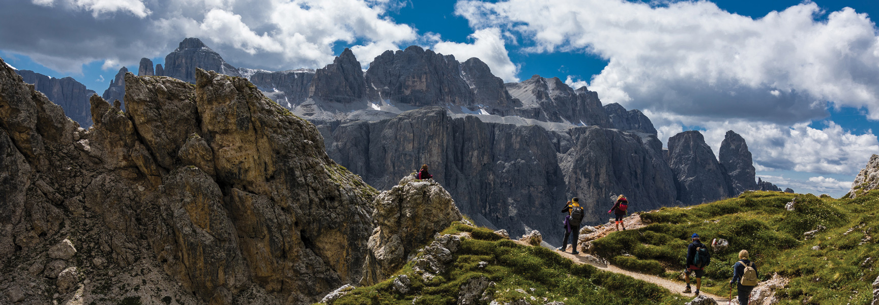 Hikers trek on a grassy path in front of a dramatic, rocky mountain range in Austria, Germany, or France.