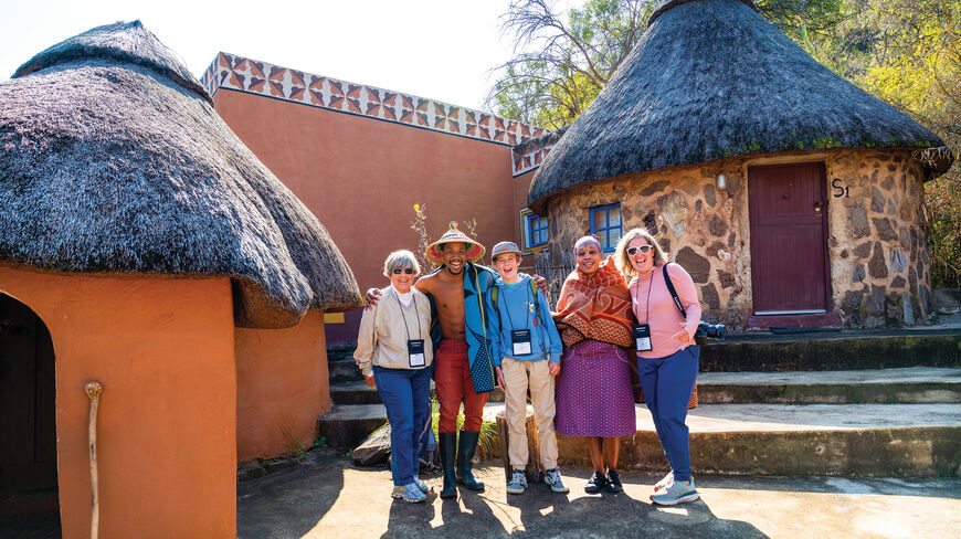A family on an African safari stands with local hosts in front of traditional thatched-roof buildings in South Africa.