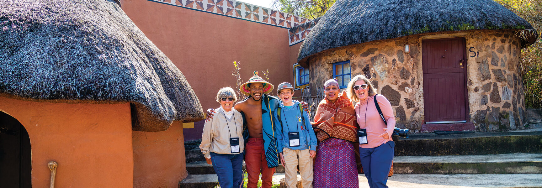 A family on an African safari stands with local hosts in front of traditional thatched-roof buildings in South Africa.