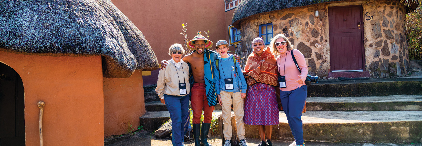 A smiling family on safari poses with local guides in front of traditional homes in South Africa.