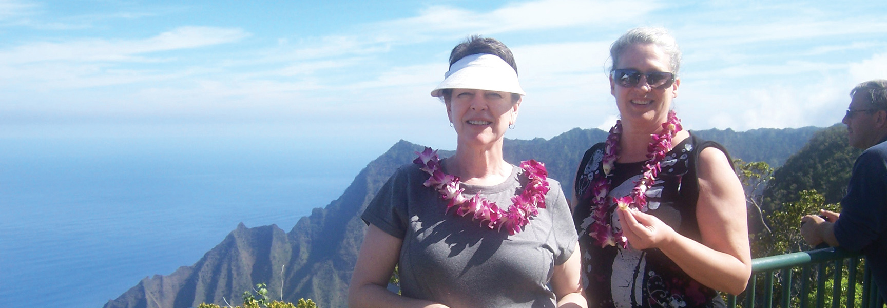 Two smiling women wearing flower leis stand at a scenic overlook with a mountainous coastal view in Hawaii.