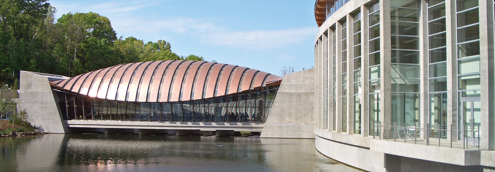The modern, copper-roofed Crystal Bridges Museum of American Art is surrounded by water and lush green trees in Arkansas.