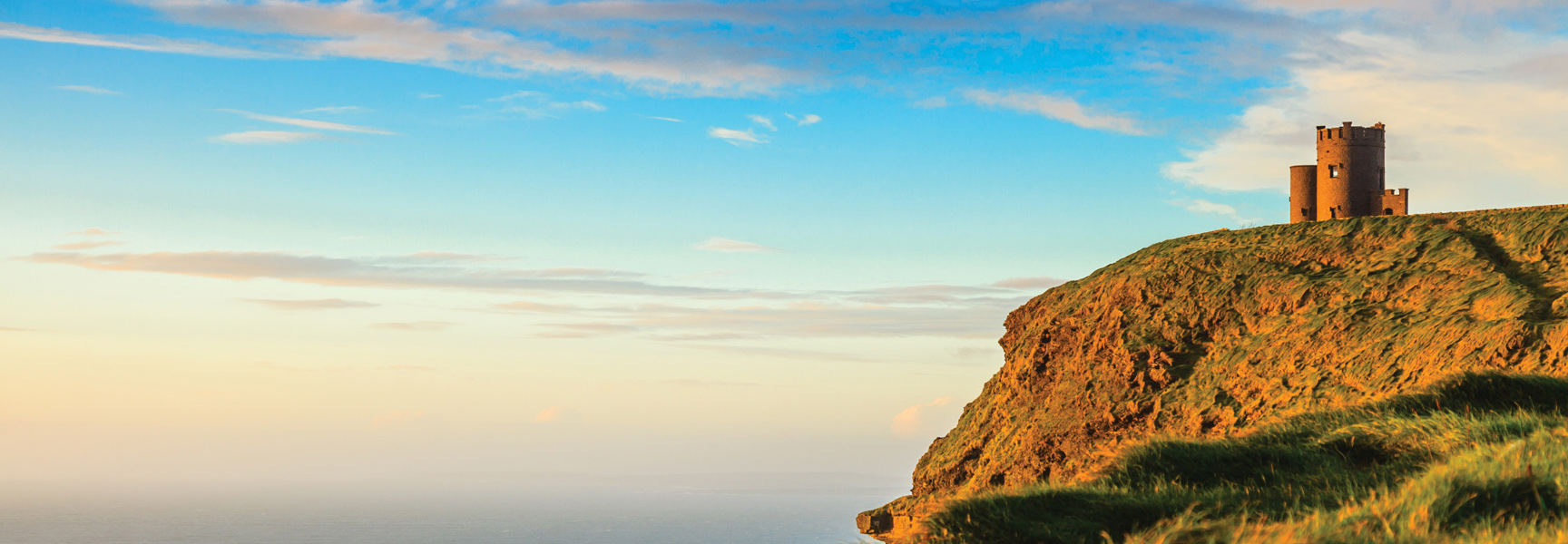 A stone tower sits on a grassy cliff overlooking the ocean in Ireland during a golden sunset.