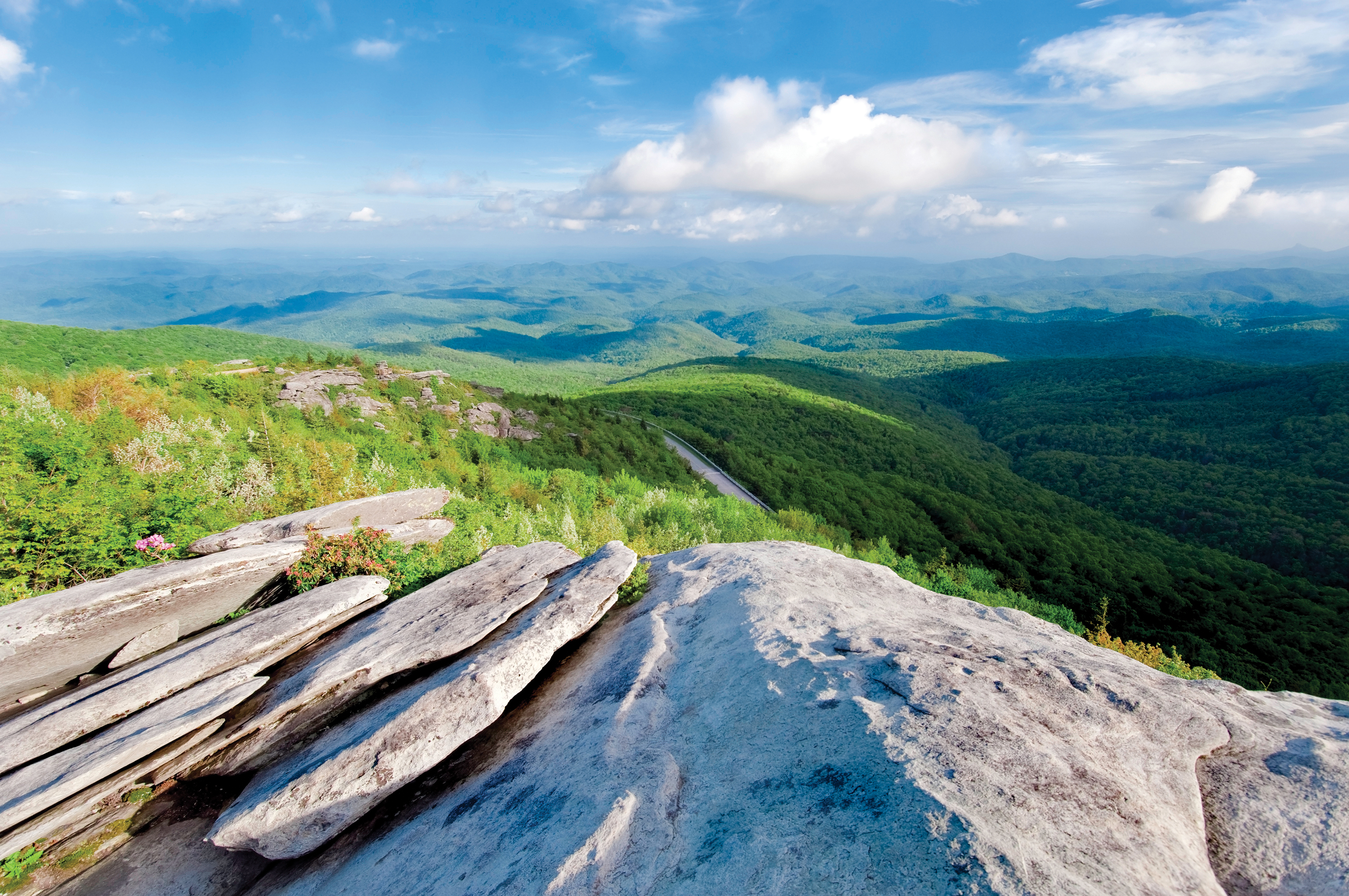 A vast view of the lush, green Southern Appalachian Mountains in Tennessee seen from a rocky overlook under a blue, cloudy sky.