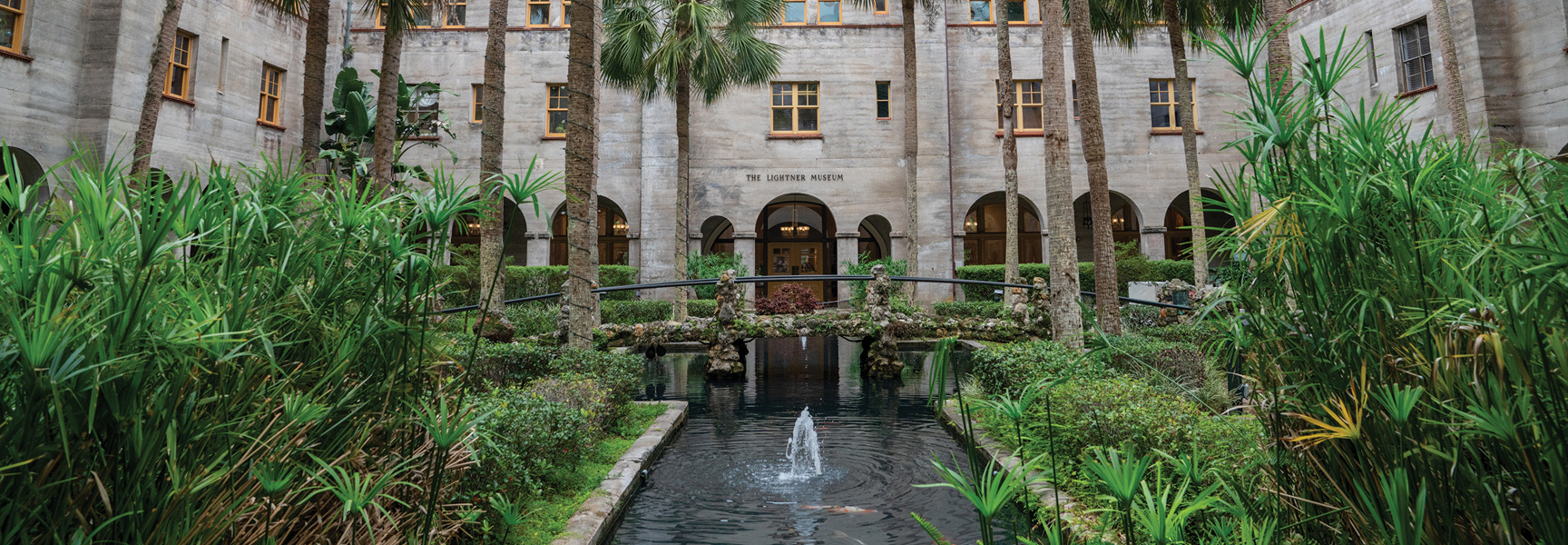The Lightner Museum in St. Augustine, Florida, seen from across a lush courtyard with a fountain, a pond, and a stone bridge.
