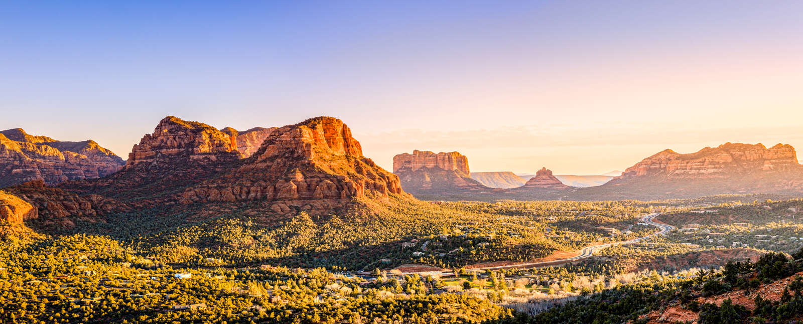 A panoramic view of red rock mountains and a winding road through the lush desert landscape of Arizona under a warm sunset sky.