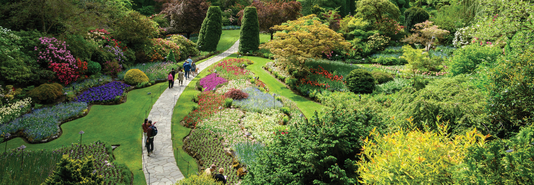 Visitors walk along a winding path through a lush, sunken garden filled with vibrant, colorful flowers and trees in British Columbia.