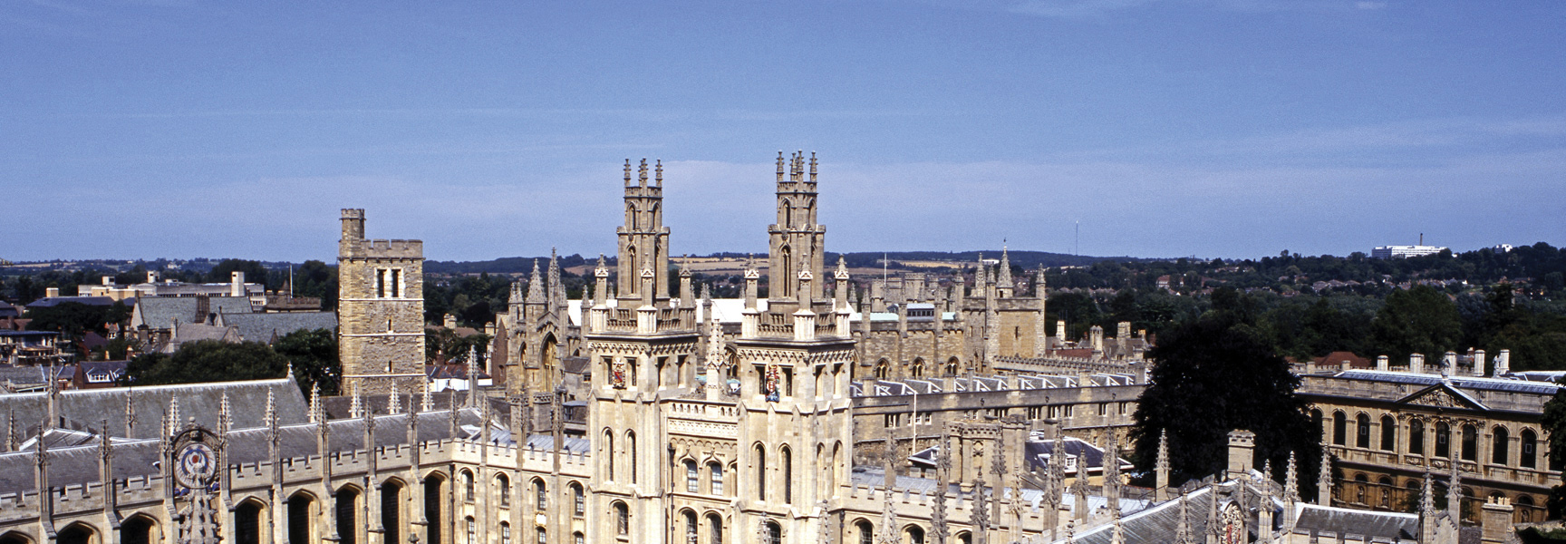 An aerial view of the historic stone towers and rooftops of Oxford University in England under a clear blue sky.
