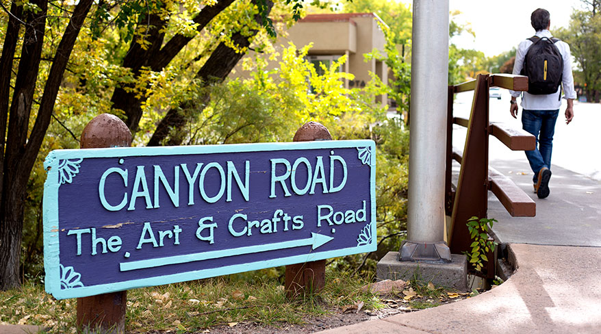A sign for Canyon Road, The Art &amp; Crafts Road, stands in Santa Fe, New Mexico, near trees and a person walking.