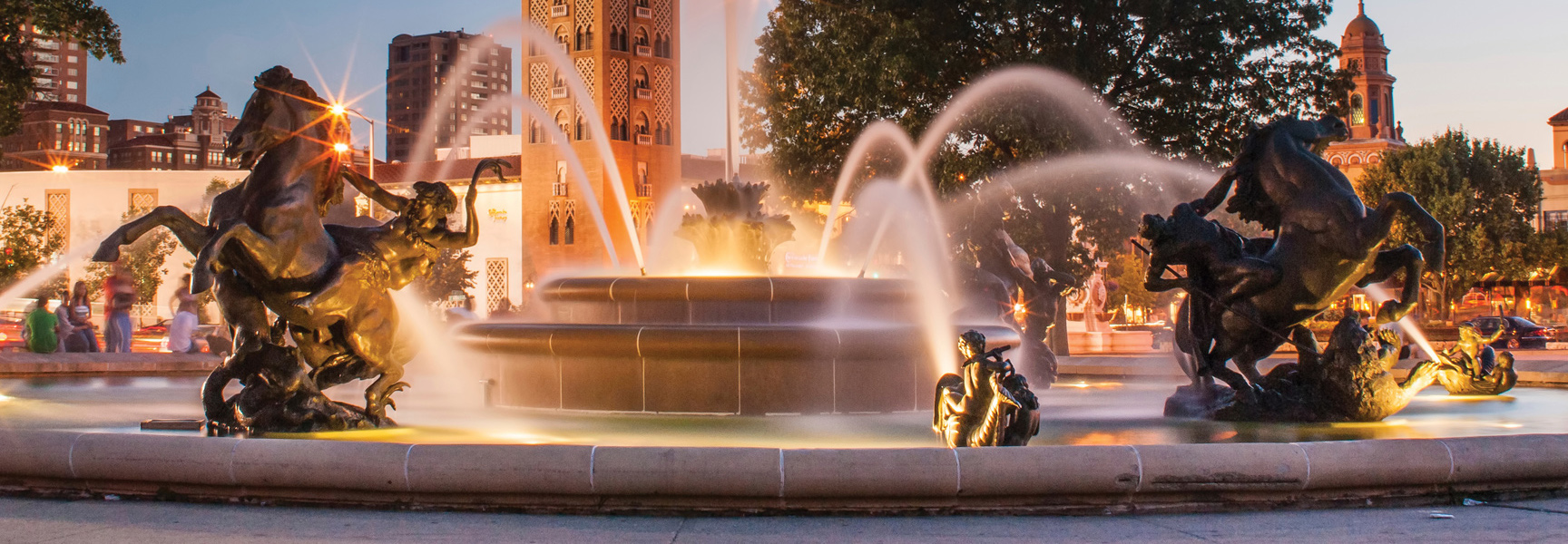 The Mill Creek Park fountain in Kansas City, Missouri, is illuminated at dusk, showcasing its large bronze equestrian statues and flowing water.