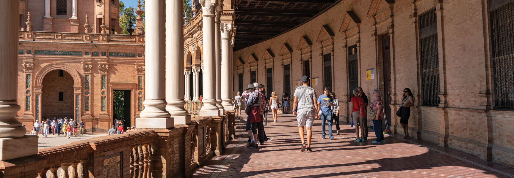 People walk through a long, arched gallery with white columns and tiled floors, showcasing the unique architecture of Sevilla, Spain.