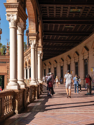 People walk through a long, arched gallery with white columns and tiled floors, showcasing the unique architecture of Sevilla, Spain.