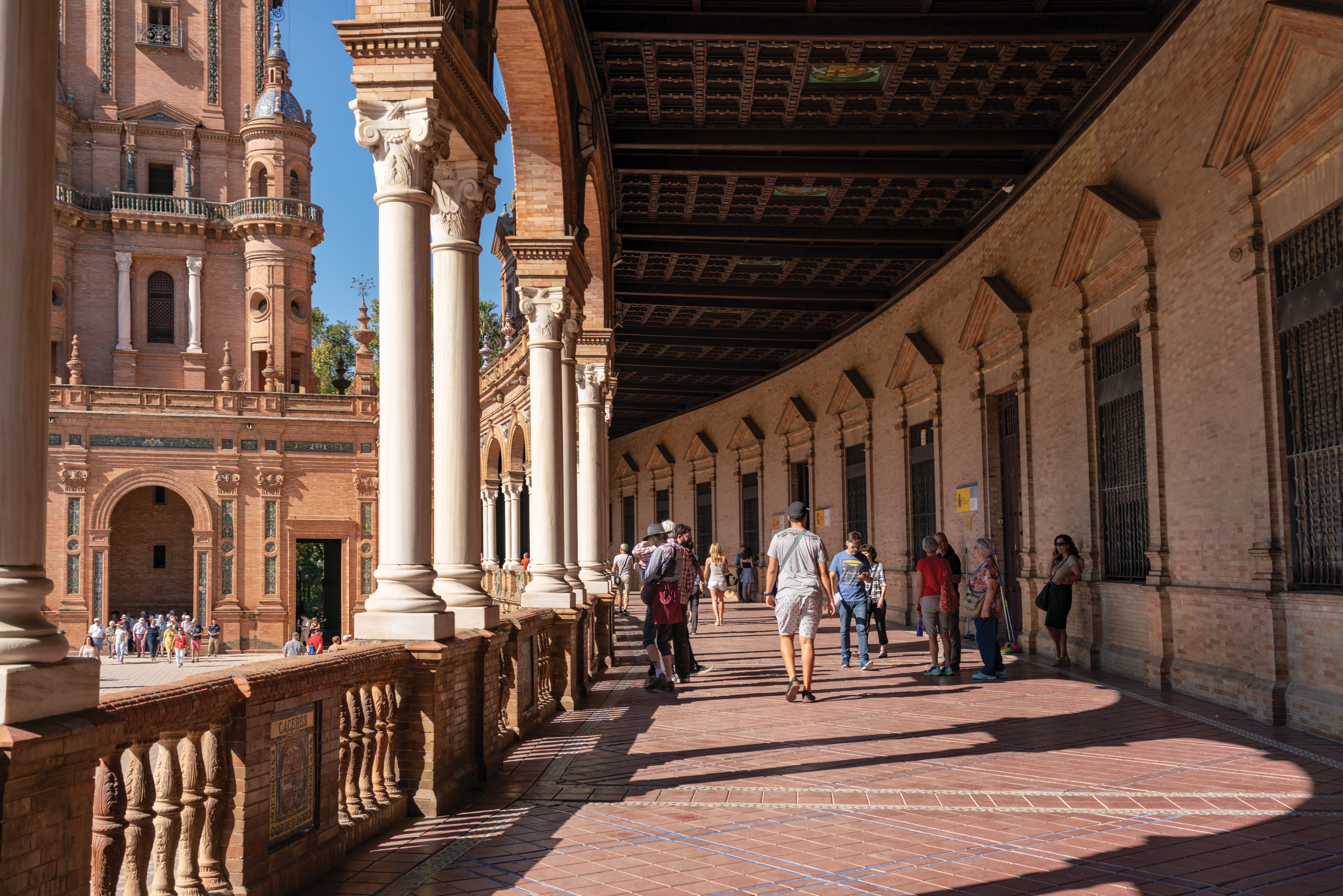 People walk through a long, arched gallery with white columns and tiled floors, showcasing the unique architecture of Sevilla, Spain.