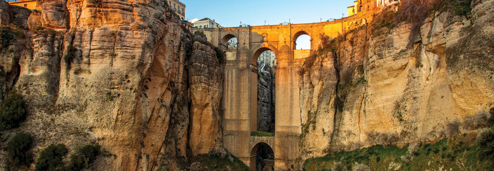 The historic, multi-arched stone bridge in Ronda, Spain, spans a deep gorge, glowing in the warm light of sunset.
