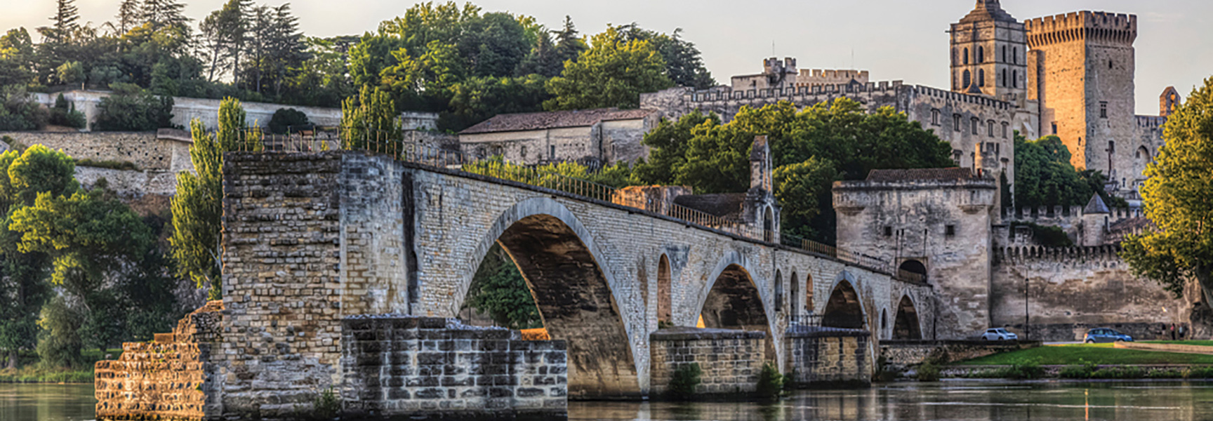 A historic stone bridge with large arches crosses a river in front of a large palace and lush trees in France.