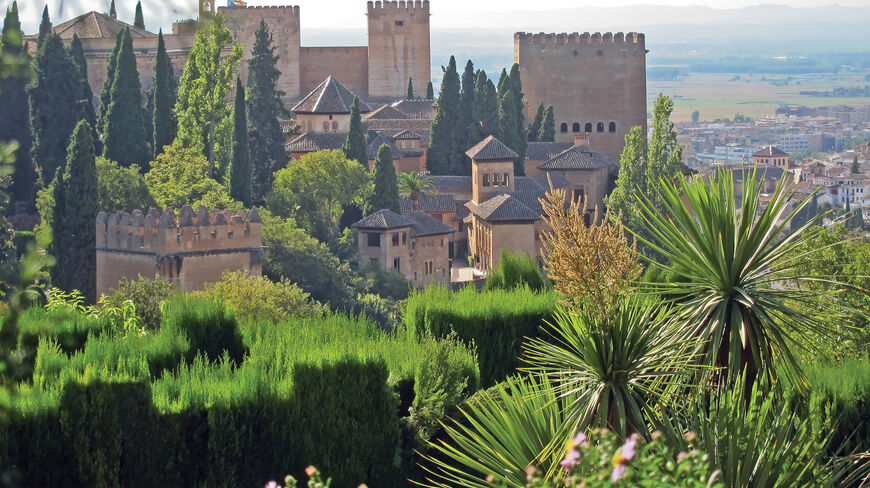 The historic Alhambra fortress and palace complex in Andalusia, Spain, surrounded by lush green trees and gardens overlooking a valley.
