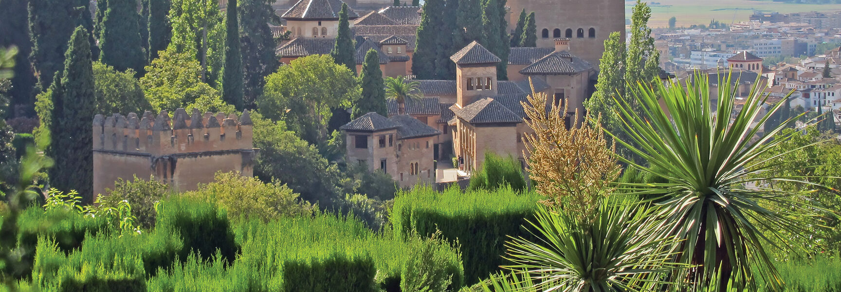 The historic Alhambra fortress and palace complex in Andalusia, Spain, surrounded by lush green trees and gardens overlooking a valley.