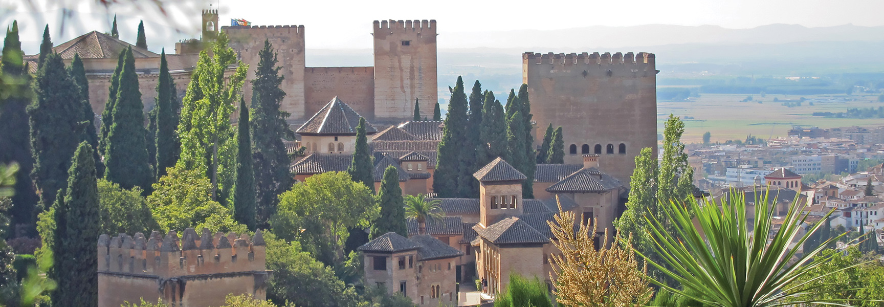 A sprawling stone fortress with towers stands among lush green trees overlooking a city in Andalusia, Spain.