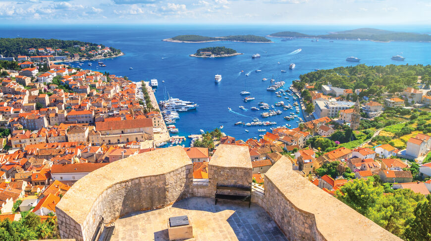 High-angle view from a stone fortification overlooking a Croatian coastal town with red-tiled roofs, a busy harbor, and islands in the sparkling Adriatic Sea.